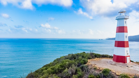 Red and white lighthouse is on a rocky cliff overlooking the ocean. The lighthouse is tall and has a...
