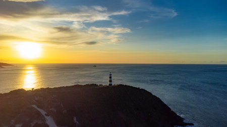 Lighthouse is on a hill overlooking the ocean. The sun is setting in the background. The sky is cloudy and the water is calmの写真素材