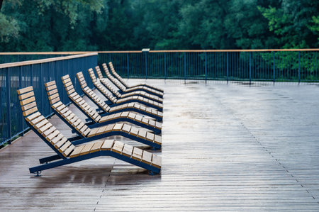 Empty wooden lounge chairs line a wet outdoor deck after rain, set against a lush green park backdrop. The scene is serene and peaceful, capturing stillness after a downpour.の写真素材