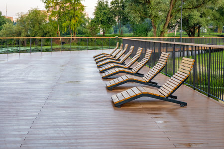 Empty wooden lounge chairs line a wet outdoor deck after rain, set against a lush green park backdrop. The scene is serene and peaceful, capturing stillness after a downpour.の写真素材