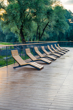 Empty wooden lounge chairs line a wet outdoor deck after rain, set against a lush green park backdrop. The scene is serene and peaceful, capturing stillness after a downpour.の写真素材