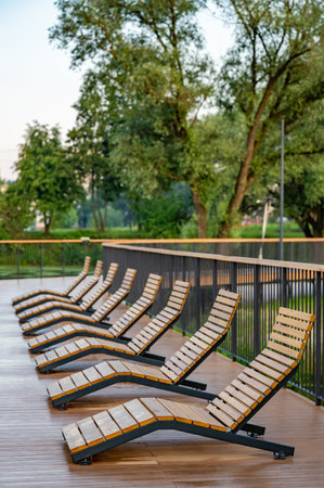 Empty wooden lounge chairs line a wet outdoor deck after rain, set against a lush green park backdrop. The scene is serene and peaceful, capturing stillness after a downpour.の写真素材