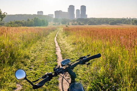 View from a bicycle handlebar on a scenic morning ride through the countryside. Sunlight illuminates the path surrounded by tall grass, with city buildings in the distance.の写真素材