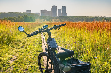 View from a bicycle handlebar on a scenic morning ride through the countryside. Sunlight illuminates the path surrounded by tall grass, with city buildings in the distance.の写真素材