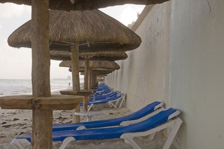 A column of straw and bamboo umbrellas into the distance along blue chaise lounge chairs on the beachの写真素材