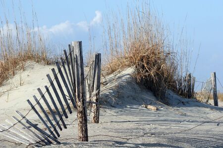 A broken fence running through sand dunes and sea oats against a blue skyの写真素材