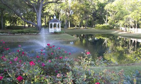 A gazebo on the shore of a lake with a fountain across from beautiful rose bushesの写真素材