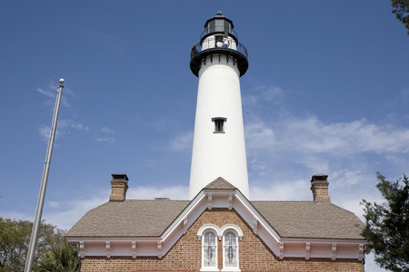 A white lighthouse rising into the blue sky behind brick house and roofの写真素材