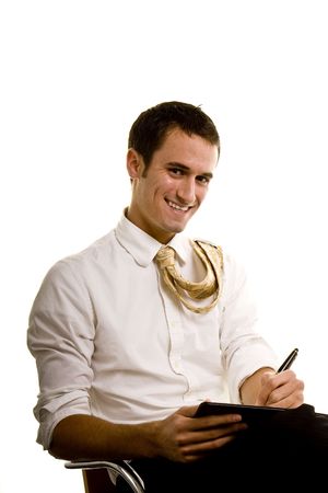 Young man wearing dress shirt and tie, sitting in a chair writing in a notebook smiling at the cameraの写真素材