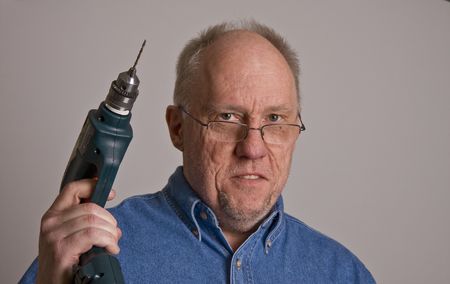 An older bald man in a blue denim shirt on a grey background wearing glasses and holding a power drillの写真素材