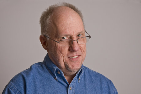 An older bald man in a blue denim shirt on a grey background smiling