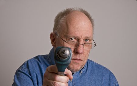 An older bald man in a blue denim shirt on a grey background wearing glasses and holding a power drillの写真素材