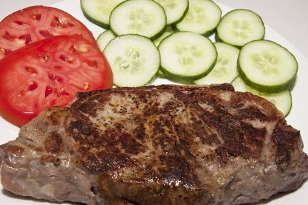 A nice strip steak on a white plate with sliced tomatoes and cucumbersの写真素材