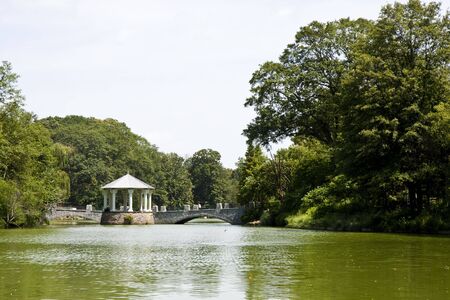 A stone gazebo and walkway across a calm green lakeの写真素材