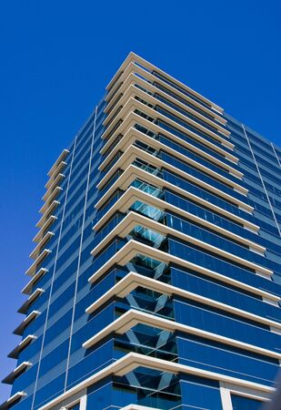 A modern blue glass office building with white corners into a blue sky.の写真素材
