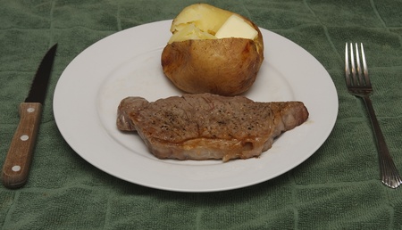 A grilled strip steak and hot buttered baked potato on a white plate with knife and fork on a green placematの写真素材