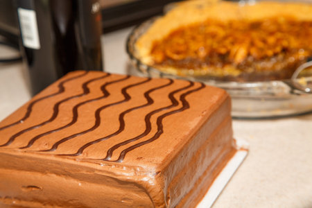 Iced chocolate cake on kitchen counter with a cut pecan pie and bottle of wine in the background