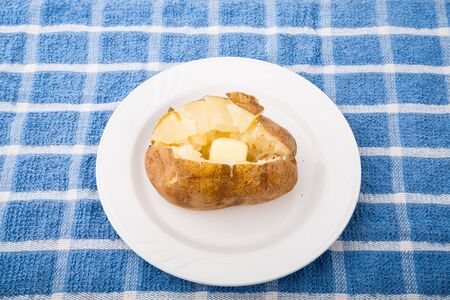 A hot, buttered, baked potato on a white plate on a blue plaid towelの写真素材