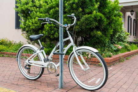 An old white bicycle locked to a sign post on a brick sidewalk in front of a green shrub and home
