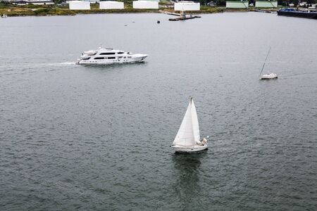 White Sailboats and Luxury Yacht in Calm Blue Bay past white fuel tanksの写真素材