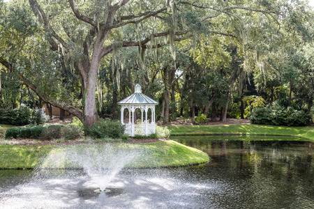 A white gazebo by an old oak tree draped in spanish moss with a fountain and lakeの写真素材