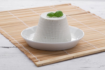 fresh ricotta cheese on white plate with basil leaf resting on wooden placemat on white wooden tableの写真素材