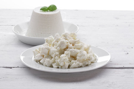 fresh ricotta cheese on white plate with basil leaf and crushed ricotta cheese on white plate all set on rustic white wooden tableの写真素材