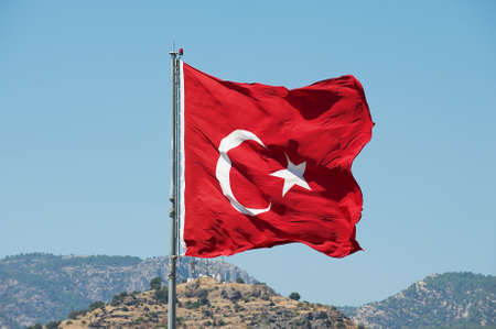 Turkish National flag at the flagpole waves over a hill with a blue summer sky at the background in...