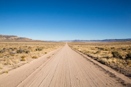 A long, straight dirt road cuts through the sage brush and empty desert of Western Utah. A low, craggy mountain can be seen on the left and a few far off peaks on the right, but otherwise a rather flat, isolated feeling.の写真素材