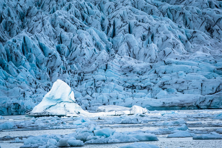 A large iceberg floating in the glacial laggon of Jokulsarlon, Iceland, is lit by the sun while the foreground icebergs and background glacier are all shadowed by cloudsの写真素材