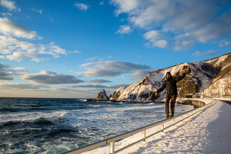 Walking along a rail in the snow, a young man balances next to the highway overlooking a rocky beach on the coast of Hokkaido, Japan.の写真素材