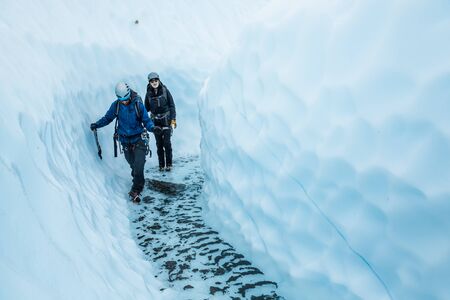 Young man and woman walking through a canyon of glacier ice on the Matanuska Glacier. In the bottom of the canyon is clear, cold water deep enough to soak their boots if they're not careful.の写真素材
