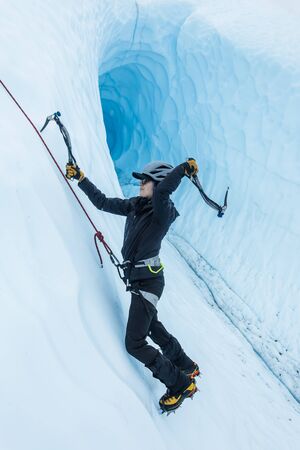 Ice climber in front of a massive ice cave in Alaska. She swings an ice tool behind her head as she...