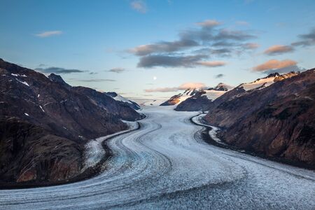 The full moon lowers in the sky during sunrise over the Salmon Glacier. Clouds turn orange in the...