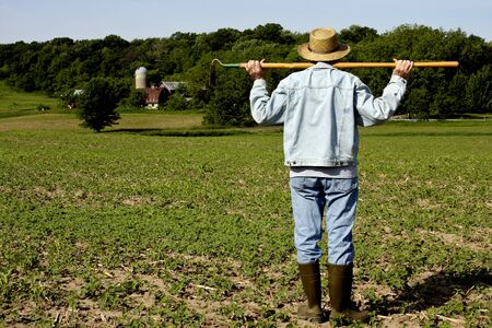 farmer standing in a field with a hoe on his shoulders with trees in the backgroundの写真素材