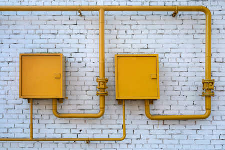 Yellow gas pipes with two closed boxes on a white brick wallの写真素材