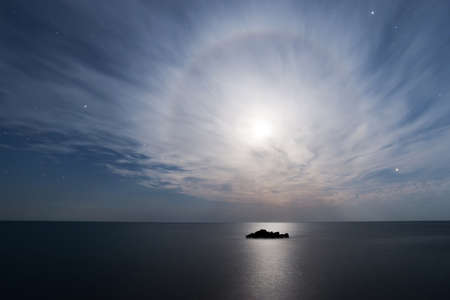 Full moon halo in the starry sky above the sea with a small rockの写真素材