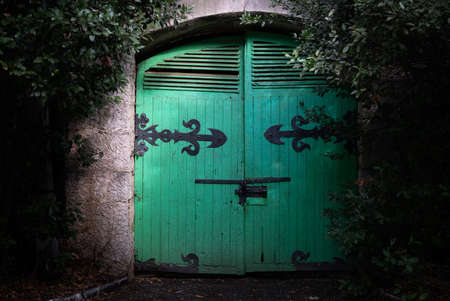The old green painted wooden gates on black wrought iron hinges in the stone wall under the treesの写真素材
