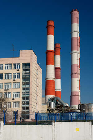 CHP station with tall red and white pipes without smoke behind a fence with barbed wire and an empty yellow sign on a blue sky backgroundの写真素材