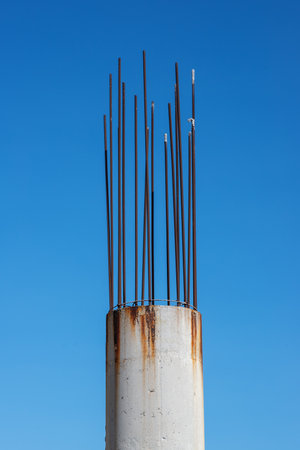Unfinished and abandoned round reinforced concrete column with protruding rusty rebar on a blue sky...