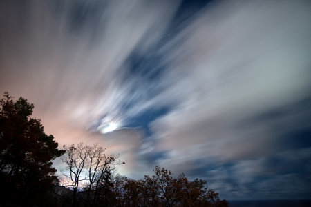 The movement of clouds in the night moonlit sky on a long exposure above the forest silhouetteの写真素材