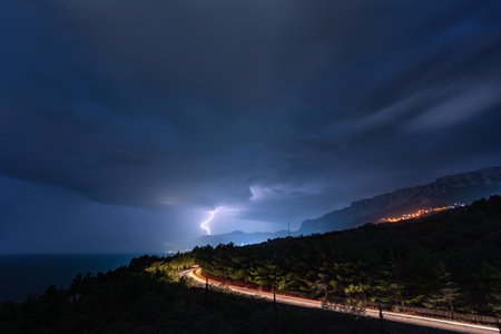 Night thunderstorm over the southern coast of Crimea, distant lightning over the mountains and the road with the light trail of a car in the foregroundの写真素材