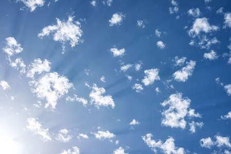 White summer clouds on a blue sky with the sun in the lower left corner.の写真素材