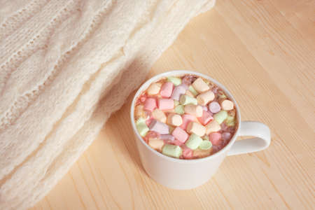 Hot chocolate beverage with colorful marshmallows on light wooden background with white fluffy knitting blanket. Hygge style coziness conceptの写真素材