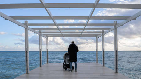 Young father walking with stroller on the pier with blue sea on background. Concept of lonely father or parenting alone.の写真素材