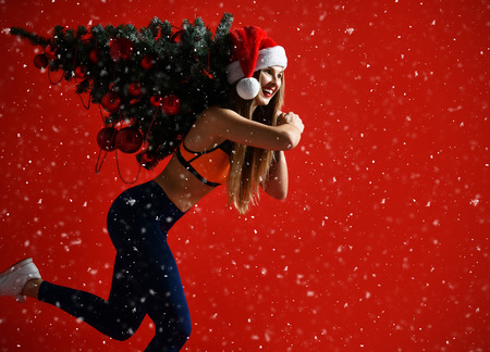 sexy Christmas fitness sport woman wearing santa hat holding  xmas tree on her shoulders. running forward on a snowing red backgroundの写真素材