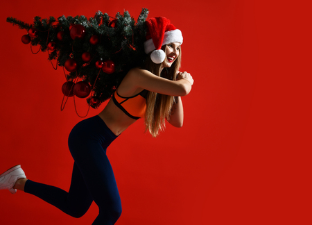 sexy Christmas fitness sport woman wearing santa hat holding  xmas tree on her shoulders. running forward on a red backgroundの写真素材