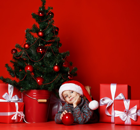 Funny smiling childboy in Santa red hat lying on  Christmas tree  holds in one hand a Christmas ball toy  on red backgroundの写真素材