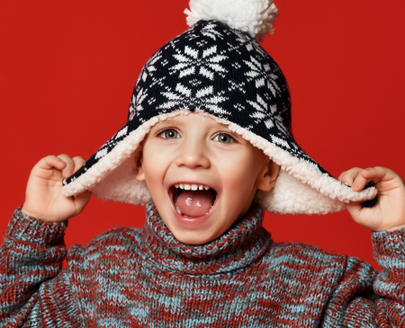 Happy child boy toddler in knitted hat and sweater having fun screaming laughing with open mouth close up winter composition over red background. December christmas conceptの写真素材