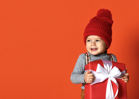 Child baby toddler kid in knitted red hat with pompon, hold big handmade present gift box isolated on a orange backgroundの写真素材
